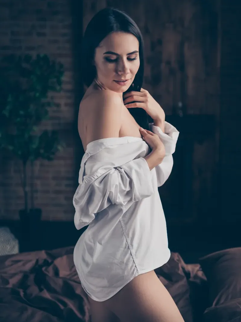 Hôtesse en chemise blanche entrouverte dans une lumière tamisée, prête pour la cam